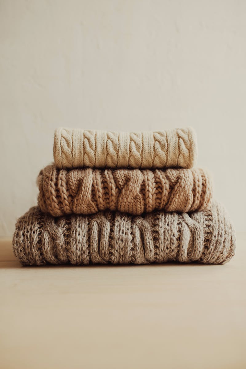 Stack of cable-knit sweaters in cream, camel, and natural tones
